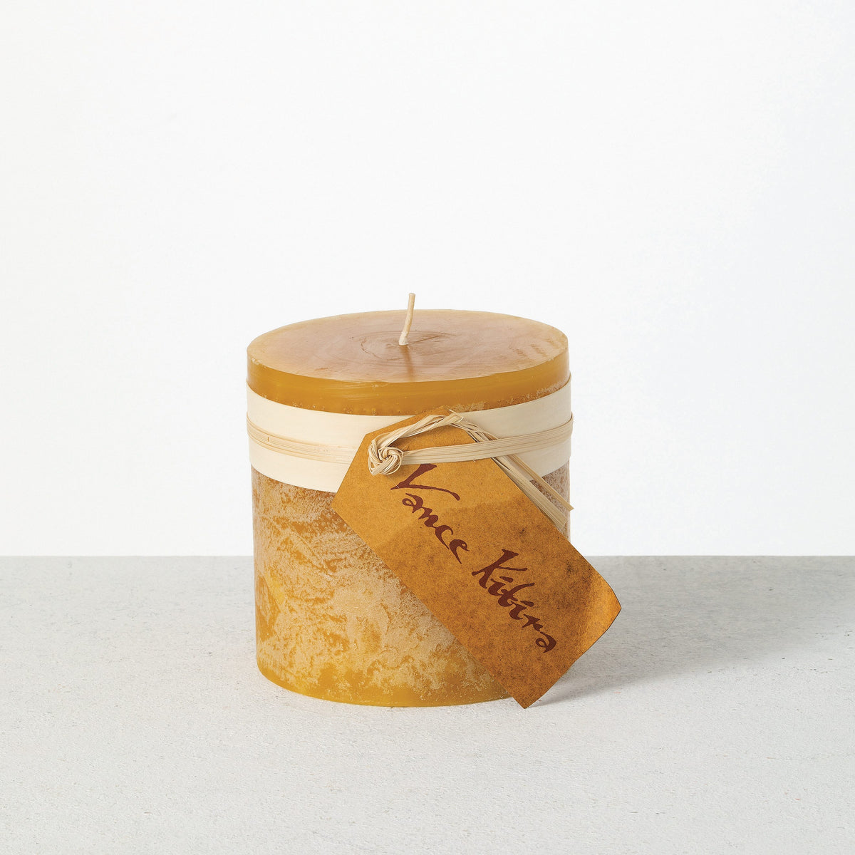 Timber Pillar Candle 4 x 4 Inch, A wide short pillar candle in golden brown sits with a cream paper band and a hang tag around its middle.