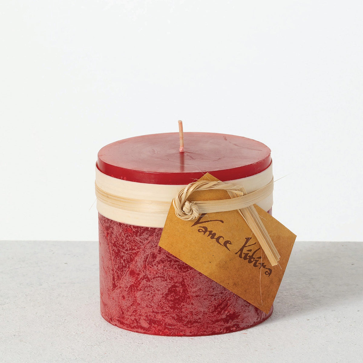Timber Pillar Candle 4 x 4 Inch, A wide short pillar candle in golden brown sits with a cream paper band and a hang tag around its middle.