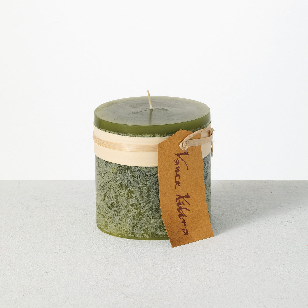 Timber Pillar Candle 4 x 4 Inch, A wide short pillar candle in golden brown sits with a cream paper band and a hang tag around its middle.
