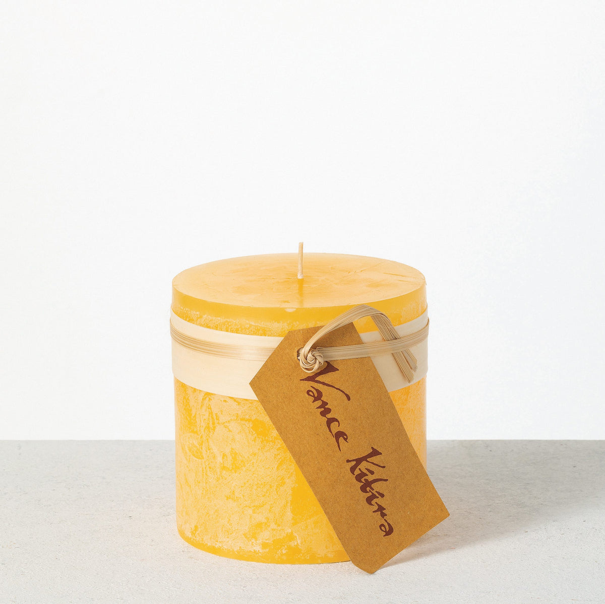 Timber Pillar Candle 4 x 4 Inch, A wide short pillar candle in golden brown sits with a cream paper band and a hang tag around its middle.