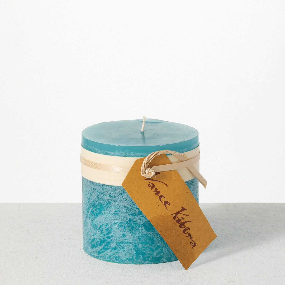 Timber Pillar Candle 4 x 4 Inch, A wide short pillar candle in golden brown sits with a cream paper band and a hang tag around its middle.