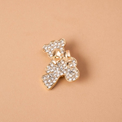Timeless Brooch, A small gold brooch pin shaped like a teddy bear and covered edge to edge in clear rhinestones, with a gold bow at its neck, shown on a tan background.
