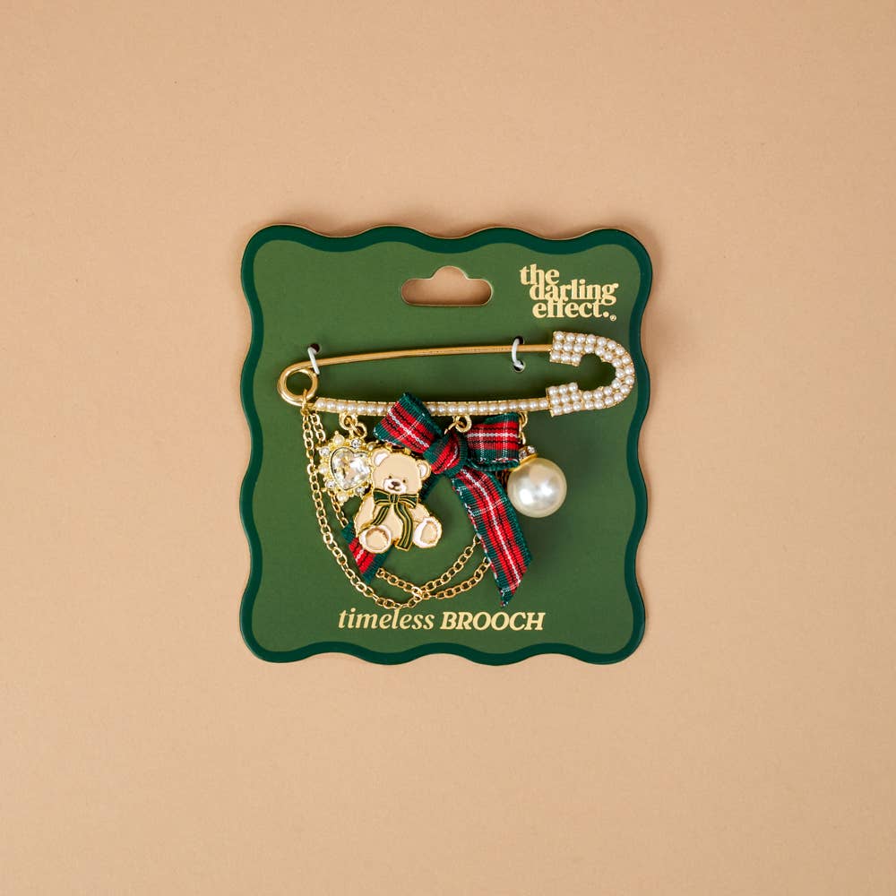 Timeless Brooch, A small gold brooch pin shaped like a teddy bear and covered edge to edge in clear rhinestones, with a gold bow at its neck, shown on a tan background.