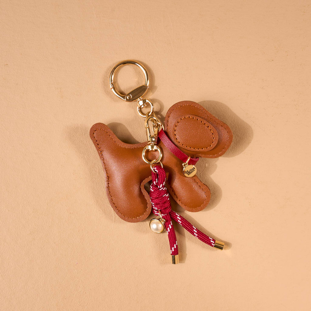 Vegan Leather Bag Charm, A dog shaped bag charm in cognac vegan leather with floppy ears, a small collar tag, and a burgundy cord tassel with a pearl bead.