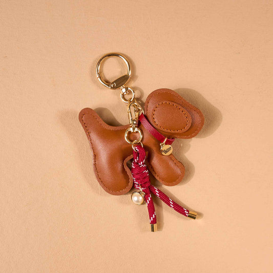 Vegan Leather Bag Charm, A dog shaped bag charm in cognac vegan leather with floppy ears, a small collar tag, and a burgundy cord tassel with a pearl bead.
