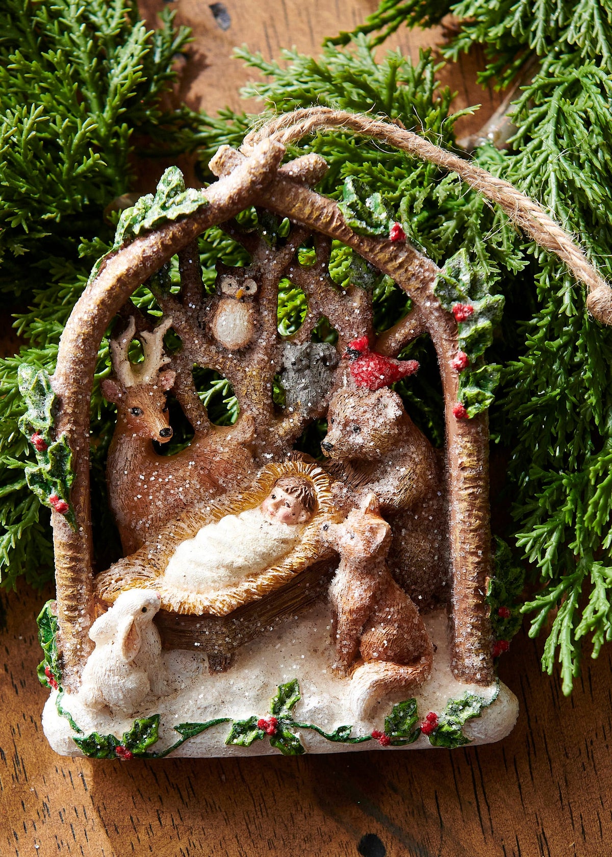 Woodland Nativity Ornament, A resin ornament shaped like an arched branch frame holding the Nativity manger scene surrounded by a deer, owl, fox, rabbit, bear, and cardinal, dusted with glitter snow.