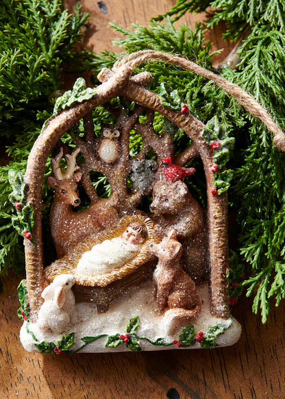 Woodland Nativity Ornament, A resin ornament shaped like an arched branch frame holding the Nativity manger scene surrounded by a deer, owl, fox, rabbit, bear, and cardinal, dusted with glitter snow.