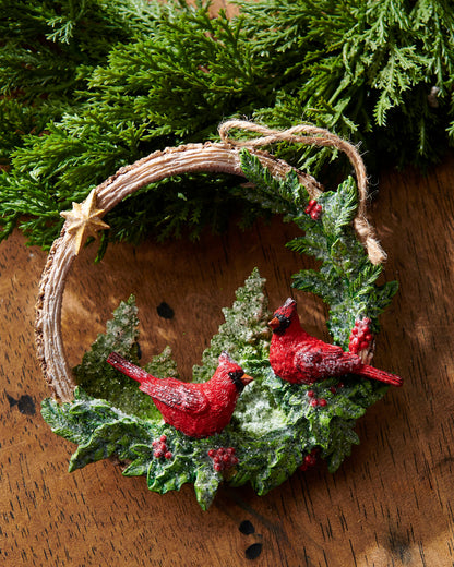 Woodland Noel Cardinals Ornament, A resin wreath ornament with two red cardinals, snow flocked pine boughs, holly berries, and a gold star, hanging from a jute cord.